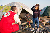 Rotkreuzhelfer mit roter Einsatzjacke kümmert sich um zwei kleine Kinder vor einem Zelt